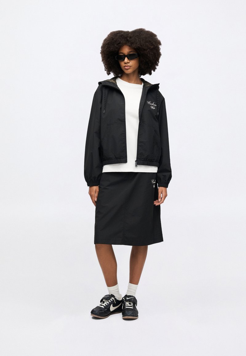 Black lightweight jacket with a hood, paired with a white top and black skirt. Featuring sneakers and sunglasses for a casual look.