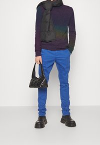 Neulepusero, jossa on violetti ja sininen liukuväri, siniset housut, musta tikattu huivi, musta tikattu käsilaukku, kömpelöt mustat saappaat.