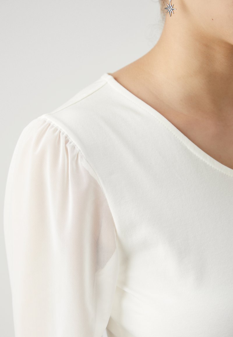 Witte top met een stevig lijfje en doorschijnende pofmouwen. De halslijn heeft een zachte v-vorm, voorzien van een gladde textuur en subtiele glans.