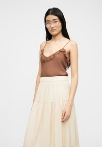 Γυναίκα με μακριά σκούρα μαλλιά, που φορά διαφανή γυαλιά, καφέ τοπ χωρίς μανίκια με βολάν και κρεμ φούστα με στρώσεις, πάνω σε απλό φόντο.