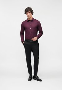 Jeune homme debout portant un pantalon noir, des chaussures noires et une chemise bordeaux à manches longues boutonnée, sur un fond blanc uni.