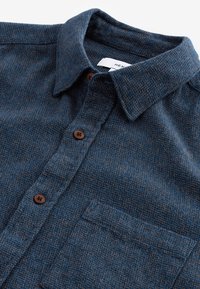 Camisa de franela azul marino con un tejido texturizado, cuello abotonado, botones marrones y un bolsillo en el pecho. Etiqueta visible dentro del cuello.