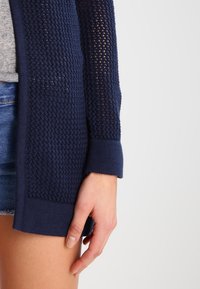 Granatowy dzianinowy kardigan o otwartej fakturze dziania, z prążkowanymi mankietami i otwartym przodem, w zestawieniu z szarą koszulką i dżinsowymi szortami.