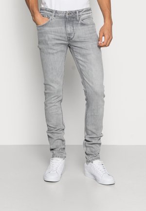 Vaqueros de mezclilla gris claro, corte ajustado, cinco bolsillos, ligero desgaste, trabillas para cinturón, combinados con zapatillas blancas sobre un fondo neutro.