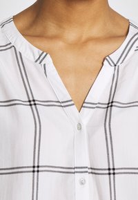 Gros plan sur une personne portant une chemise blanche à rayures noires en tartan et un col rond avec des boutons sur le devant.