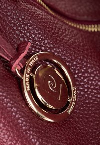 Borsa in pelle bordeaux con superficie testurizzata, caratterizzata da un ciondolo circolare dorato inciso con "MADE IN ITALY" e un design a spirale.