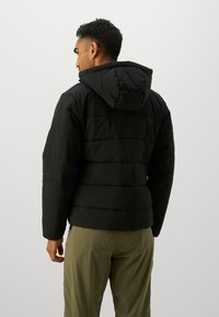 Homme portant une veste matelassée à capuche noire et un pantalon vert olive, vu de dos sur un fond clair.