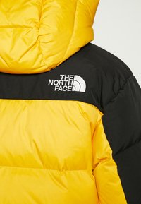 Geltonos ir juodos pūkinės striukės su siuvinėta tekstūra. „The North Face“ logotipas baltu siūlu yra siuvinėtas juodoje dalyje.