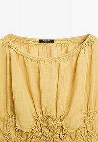 Mosterdgele blouse met een ronde halslijn, geplooid stof en decoratieve smockingdetails aan de zoom. Label zichtbaar bij de hals.