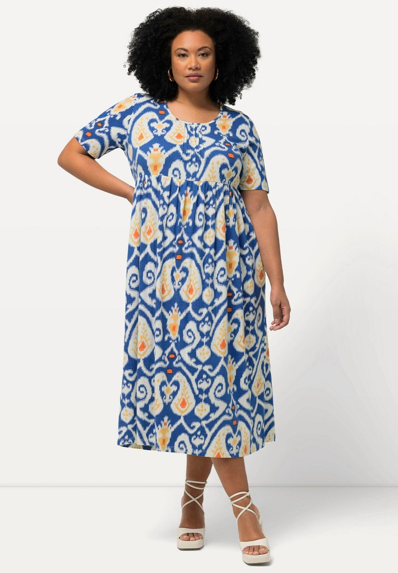 Sininen maksimekko, jossa on textured paisley-kuvio keltaisena ja oranssina. Lyhyet hihat, pyöreä pääntie ja gatheroitu vyötäröyksityiskohta.