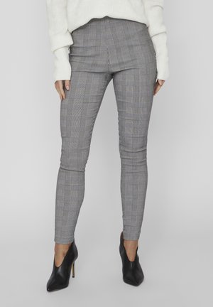 Mujer vestida con pantalones ajustados de cuadros grises, zapatos de tacón alto puntiagudos negros y un suéter blanco, posando contra un fondo liso.