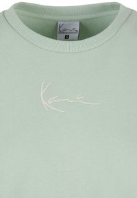 Sweat-shirt vert clair avec col ras du cou côtelé et logo "Kani" blanc brodé centré sous l'encolure. Étiquette de taille visible à l'intérieur du col.