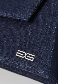 Nærbillede af mørk denimstof med et sølvmetallic logo, der viser stiliserede bogstaver "DG" på en foldet kant. Struktur synlig.