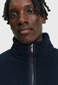Gros plan d'une personne aux cheveux courts torsadés portant une veste polaire bleu foncé avec une fermeture éclair argentée entièrement fermée.