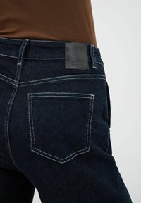 Ciemnoniebieskie dżinsy o teksturowanej tkaninie, białe przeszycia i czarna skórzana naszywka na pasku z logo marki.