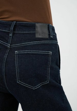 Jeans de mezclilla oscura con una tela texturizada, costuras blancas y un parche de cuero negro en la cintura con el logo de la marca.