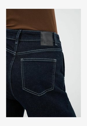 Dunkelblaue Denim-Jeans aus strukturiertem Stoff, mit weißen Nähten und einem schwarzen Lederpatch am Bund, auf dem das Markenlogo abgebildet ist.