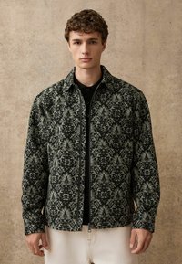 Schwarze und graue gemusterte Jacke mit Kragen, durchgehendem Reißverschluss und langen Ärmeln, über einem schwarzen Shirt getragen, präsentiert vor einem neutralen Hintergrund.