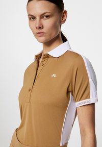 Vaaleanruskea polo-paita, jossa on valkoinen kaulus ja sivupaneelit, joissa on hienovarainen logo ja tekstuuriset yksityiskohdat hihoissa. Sileä kangaspinta.