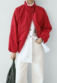 Personne portant une veste rouge courte par-dessus une chemise blanche boutonnée et un pantalon crème, tenant un sac noir dans une main et ayant l'autre main dans la poche.
