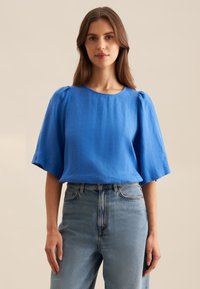 Blusa blu in lino a maniche corte con scollatura rotonda e leggermente a sbuffo, abbinata a jeans in denim chiaro a vita alta.
