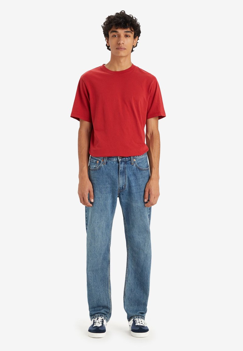 Camiseta roja de manga corta, corte relajado; jeans rectos azules con lavado claro; zapatillas casuales con cordones blancos y acentos en azul marino.