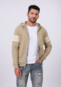 Beige zip-up hoodie met crèmekleurig accentpaneel, verstelbare trekkoorden en kangoeroezakken, gedragen over een wit t-shirt met versleten blauwe jeans.
