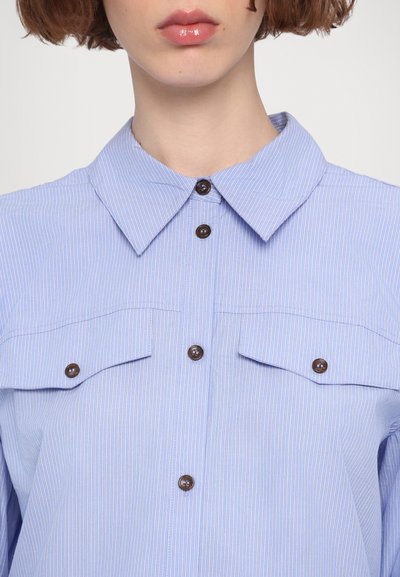 Camicia a righe azzurro chiaro con colletto button-down, due tasche sul petto e bottoni marroni. Il tessuto appare liscio con una leggera texture.