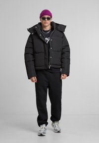 Černá péřová bunda se zipem a patentkami, vysokým límcem, v kombinaci s černými kalhotami a světle zbarvenými teniskami. Fialová čepice a sluneční brýle.