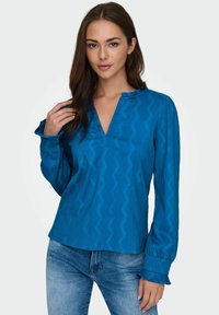 Blouse bleu à manches longues avec un décolleté en V, ornée d'un motif en zigzag et de volants sur le col et les poignets. Portée avec un jean en denim.