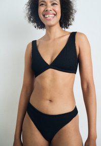 Femeie zâmbitoare cu păr creț purtând un bikini negru texturat, stând în fața unui fundal simplu gri deschis.