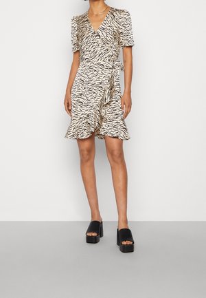 Kobieta ubrana w beżową i czarną sukienkę kopertową w paski zebry z falbaną na dole i czarne sandały na platformie z odkrytymi palcami, stojąca na białym tle.