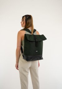 Zelený batoh s překlopnou klopou, černým dnem, nastavitelnými popruhy a přední přezkou na zapínání. Vyroben z odolné tkaniny, minimalistický design.