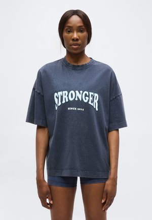 Femme portant un T-shirt oversize gris foncé avec le texte "STRONGER SINCE 2014" et un short de cycliste assorti gris foncé, debout sur fond blanc.
