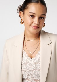 Jonge vrouw met krullend haar in een knot, draagt gouden oorringen, gelaagde gouden kettingen, een beige blazer en een witte kanten top tegen een witte achtergrond.