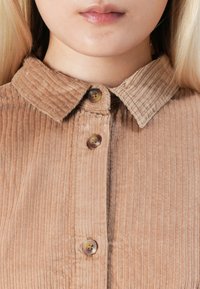 Close-up van het onderste gezicht en de nek van een persoon die een beige Corduroy shirt met drie knopen en een puntige kraag draagt.