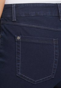 Jean en denim bleu foncé avec une poche arrière dotée d'un bouton en métal argenté et de surpiqûres blanches contrastantes. Texture lisse avec une coupe ajustée.
