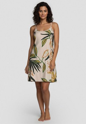 Kobieta stoi boso, ubrana w beżową sukienkę bez rękawów z zielonymi i żółtymi tropikalnymi wzorami liści na jednolitym tle.