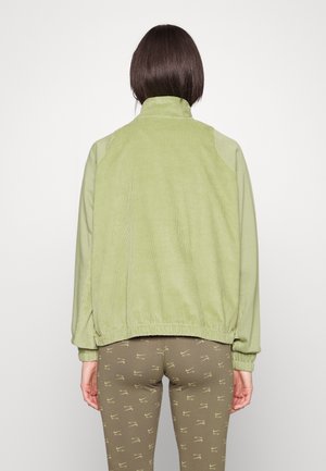 Person mit kurzen dunklen Haaren, die eine hellgrüne Jacke und olivgrüne Leggings mit wiederholten Nike Air-Logos trägt, von hinten gezeigt.
