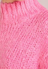 Różowy sweter wykonany na drutach z wysokim, prążkowanym kołnierzem. Tkanina wydaje się miękka i puszysta, ukazując grube, teksturowane włóczki w intensywnym odcieniu.