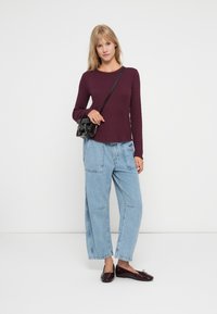 Langærmet bordeaux top, løstsiddende lyseblå denimbukser, sort crossover taske og skinnende sorte flade sko på en neutral baggrund.