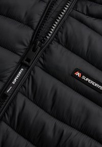 Giacca nera trapuntata con cerniera nera parzialmente aperta e una toppa con il logo Superdry visibile sul lato destro.