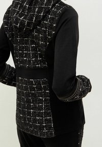 Chaqueta texturizada en blanco y negro con capucha. Presenta un patrón tejido, mangas lisas negras y costuras decorativas en los puños.