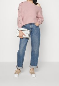 Maglione rosa off-shoulder in maglia con bottoni decorativi, jeans blu a vita alta, sneakers beige e una borsa a tracolla bianca texturizzata.