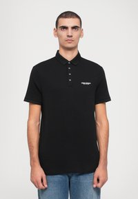 Musta polo-paita, jossa on klassinen kaulus, viiden napin lista ja lyhyet hihat. Vasemmassa rinnassa on pieni valkoinen logo.