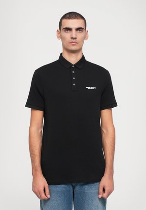 Melns polo krekls ar clásico apvalku, piecu pogu slēgšanu un īsām piedurknēm. Mazs balts logs atrodas kreisajā krūtī.