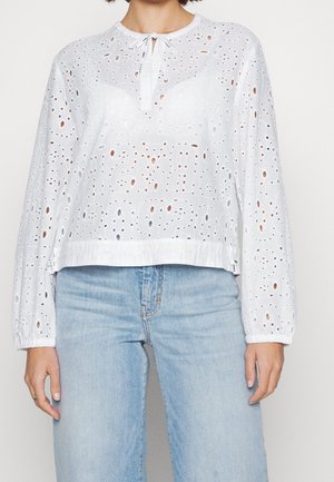 Kobieta ubrana w białą, długorękawową bluzkę z ażurowym wzorem, z małym wiązaniem z przodu oraz jasnoniebieskie jeansy z wysokim stanem.