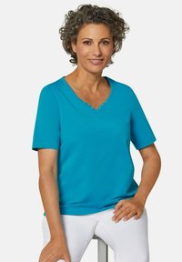 Turquoise V-hals T-shirt met korte mouwen, versierd met decoratieve studs langs de halslijn. Gladde textuur, gecombineerd met witte broek.