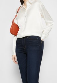 Camisa blanca de satén con botones, jeans de mezclilla azul oscuro con costuras visibles y un pequeño bolso de cuero naranja al hombro. Textura suave y diseño ajustado.