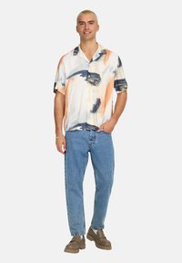 Camisa de manga corta con botones que presenta un patrón abstracto en naranja, azul y blanco. Combinada con jeans de color azul claro y zapatos marrones.